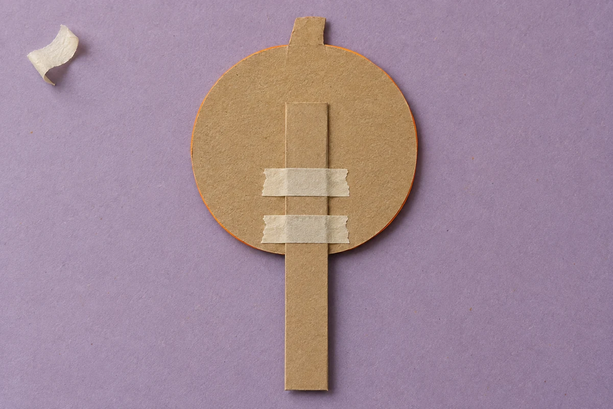 Back of a paper pumpkin puppet with a wide card handle secured by two horizontal strips of tape.